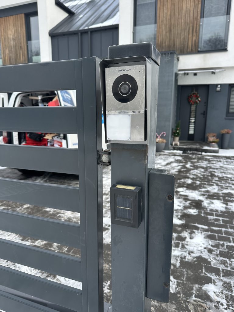 Panel wideodomofonu z kamerą na metalowym słupku przy furtce.