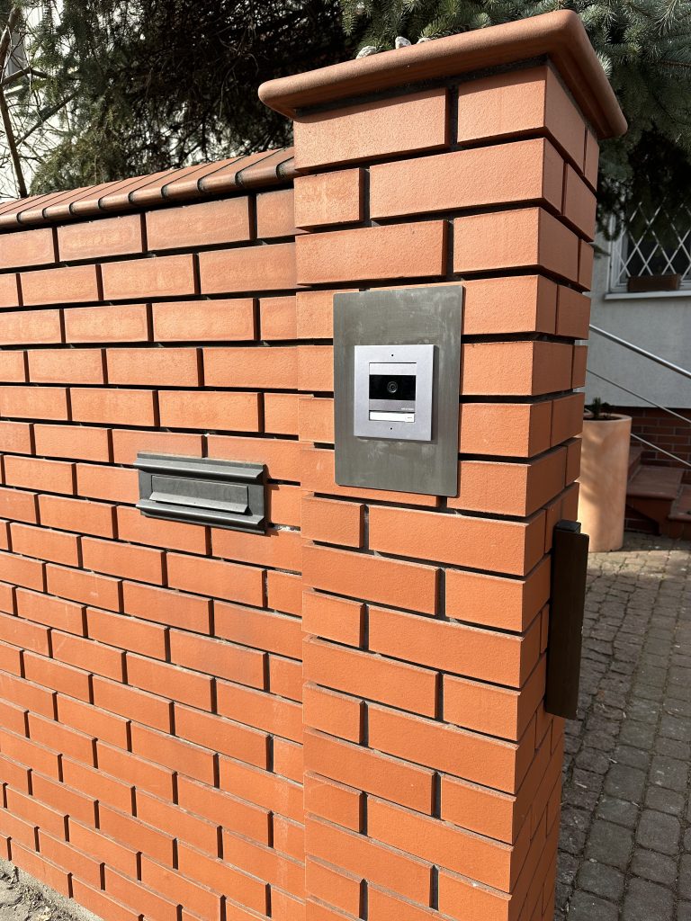 Panel wideodomofonu na metalowej płycie w słupku z czerwonej cegły.