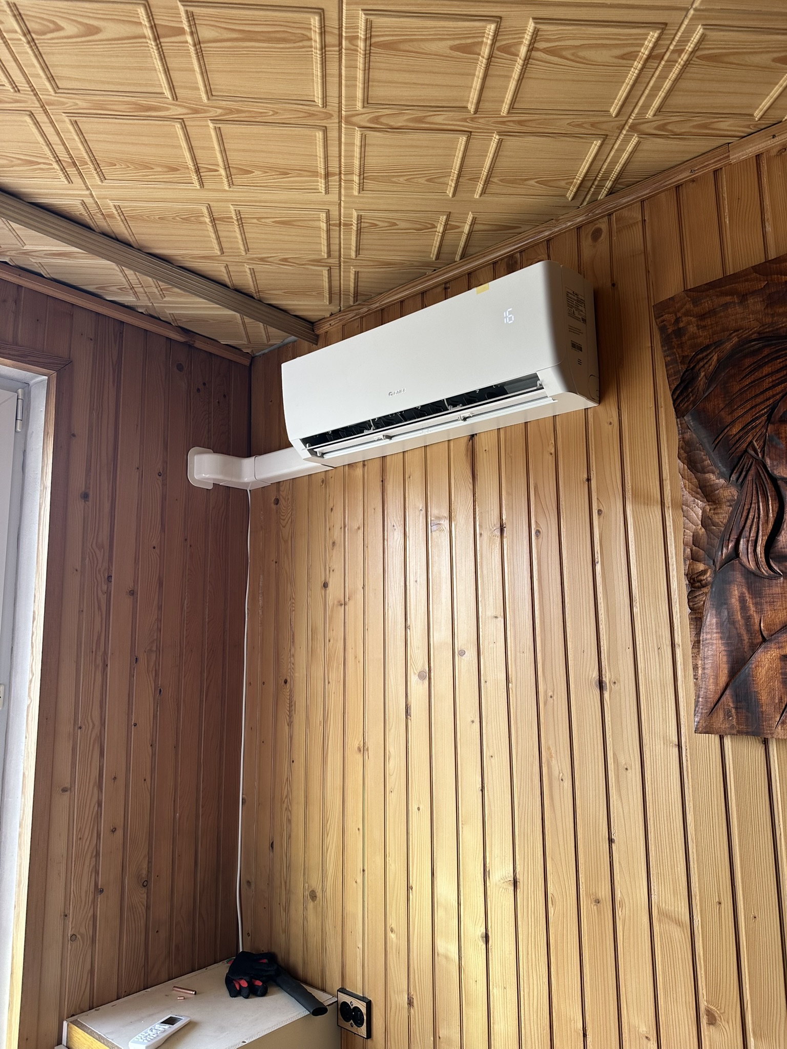 Klimatyzator ścienny z instalacją poprowadzoną w białym korytku na drewnianej ścianie.